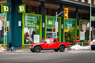 A bright red pickup truck is stopped at a crosswalk in front of a building featuring large green-framed windows. The building is branded with the TD logo and has posters with investment-themed messages. Snow is seen on the ground, and a person carrying a blue shopping bag is walking across the crosswalk.