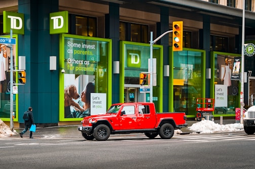 A bright red pickup truck is stopped at a crosswalk in front of a building featuring large green-framed windows. The building is branded with the TD logo and has posters with investment-themed messages. Snow is seen on the ground, and a person carrying a blue shopping bag is walking across the crosswalk.