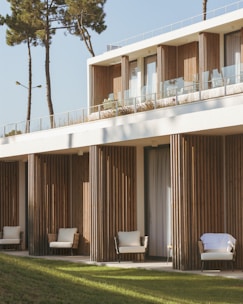 A modern building features a minimalist design with natural wooden panels and large glass windows. The structure is surrounded by green lawns and tall trees. Comfortable outdoor seating is visible on a balcony and terrace, suggesting a serene and inviting atmosphere.