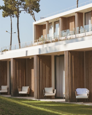 A modern building features a minimalist design with natural wooden panels and large glass windows. The structure is surrounded by green lawns and tall trees. Comfortable outdoor seating is visible on a balcony and terrace, suggesting a serene and inviting atmosphere.