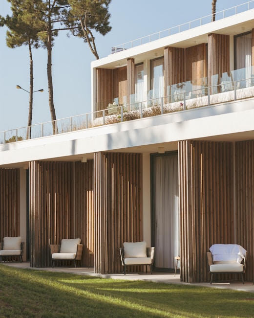 A modern building features a minimalist design with natural wooden panels and large glass windows. The structure is surrounded by green lawns and tall trees. Comfortable outdoor seating is visible on a balcony and terrace, suggesting a serene and inviting atmosphere.