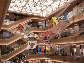 A shopping mall interior with multiple levels and escalators. Bright, geometric ceiling lights illuminate the space. A large, colorful art installation featuring cartoon-like figures is suspended in the center, adding a whimsical touch to the modern architecture.