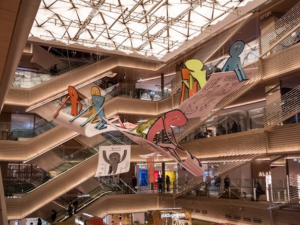 A shopping mall interior with multiple levels and escalators. Bright, geometric ceiling lights illuminate the space. A large, colorful art installation featuring cartoon-like figures is suspended in the center, adding a whimsical touch to the modern architecture.