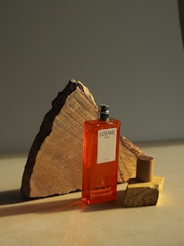 A vibrant orange perfume bottle labeled 'Loewe Solo Ella' is positioned against a background of geometric stone pieces. The composition creates a dramatic play of light and shadow.