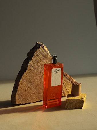 A vibrant orange perfume bottle labeled 'Loewe Solo Ella' is positioned against a background of geometric stone pieces. The composition creates a dramatic play of light and shadow.