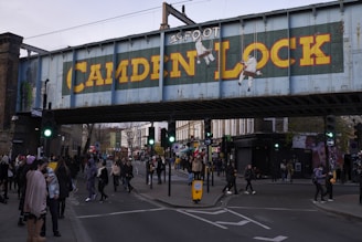 A busy urban scene features a metal bridge painted with 'Camden Lock' in bold yellow letters, surrounded by people walking through a lively street with shops and traffic lights.