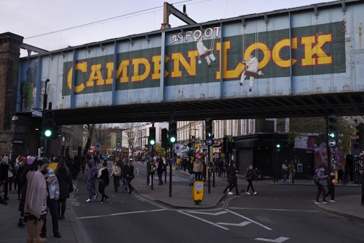 A busy urban scene features a metal bridge painted with 'Camden Lock' in bold yellow letters, surrounded by people walking through a lively street with shops and traffic lights.