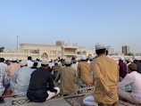 A large group of people are gathered for a prayer session outside a mosque. Most are wearing traditional attire and caps, and they are seated on patterned prayer rugs. The mosque, with its ornate design and minarets, is visible in the background under a clear blue sky. Buildings and palm trees can also be seen in the distance.
