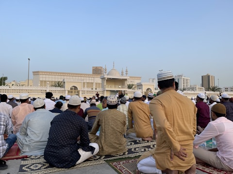 A large group of people are gathered for a prayer session outside a mosque. Most are wearing traditional attire and caps, and they are seated on patterned prayer rugs. The mosque, with its ornate design and minarets, is visible in the background under a clear blue sky. Buildings and palm trees can also be seen in the distance.