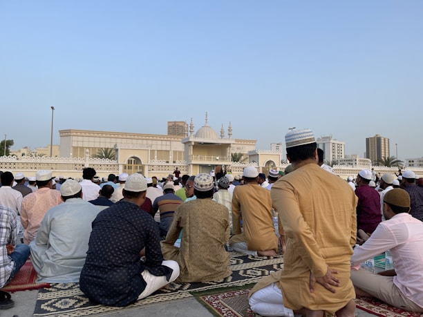 A large group of people are gathered for a prayer session outside a mosque. Most are wearing traditional attire and caps, and they are seated on patterned prayer rugs. The mosque, with its ornate design and minarets, is visible in the background under a clear blue sky. Buildings and palm trees can also be seen in the distance.
