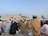 A large group of people are gathered for a prayer session outside a mosque. Most are wearing traditional attire and caps, and they are seated on patterned prayer rugs. The mosque, with its ornate design and minarets, is visible in the background under a clear blue sky. Buildings and palm trees can also be seen in the distance.