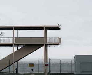 An exterior view of an elevated walkway with stairs, securely fenced off in front of a body of water. A CCTV camera is mounted on the upper level of the structure, allowing for surveillance. The overcast sky and sparse surroundings lend a minimalist feel to the scene.