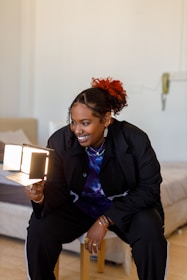 A person with a bright smile is holding a small foldable mirror with lights, sitting on a stool in a cozy room. The individual is stylishly dressed in a dark jacket and colorful tie-dye shirt, with accessories like rings and earrings. The background shows part of a bed and a softly lit wall.