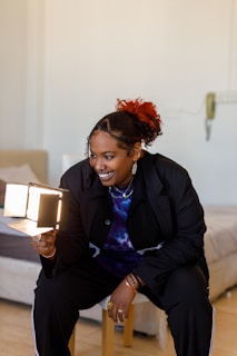 A person with a bright smile is holding a small foldable mirror with lights, sitting on a stool in a cozy room. The individual is stylishly dressed in a dark jacket and colorful tie-dye shirt, with accessories like rings and earrings. The background shows part of a bed and a softly lit wall.