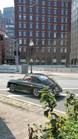 A classic black car is parked on a street corner in an urban setting. The background features tall brick buildings with multiple windows. A traffic signal is visible, as well as some greenery and yellow flowers in the foreground.