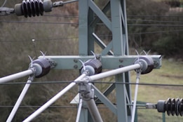 Detailed view of a metal transmission tower with several insulators and cables attached, set against a blurred natural backdrop.