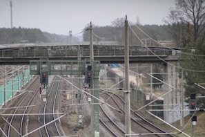 Railway tracks stretch into the distance beneath an overpass, with several power lines and signal lights visible. Surrounding the area, there are trees and a forested background, creating a rural setting.