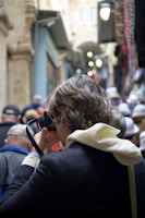 A photographer in a crowded alleyway takes a picture of something in front of them. The person has short, curly hair and wears a hoodie under a blazer while holding a camera up to their eye. The alley is busy with people wearing hats and jackets, with buildings on either side and hanging lamps lighting the way.