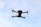 A drone is captured mid-flight against a backdrop of blue sky with scattered clouds. The drone's design appears sleek and modern, with four propellers and a camera mounted underneath.