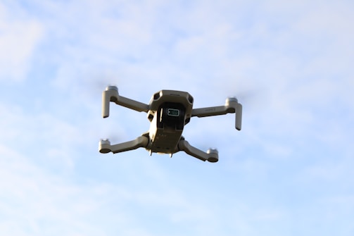 A drone is captured mid-flight against a backdrop of blue sky with scattered clouds. The drone's design appears sleek and modern, with four propellers and a camera mounted underneath.
