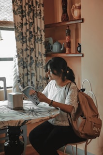 A woman sits at a table in a cozy, softly lit room with floral curtains. She is reading a book titled 'VINE' with a backpack slung over the chair. The room has warm-toned walls with shelves displaying decorative items such as teapots and vases.
