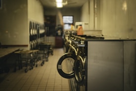 A dimly lit laundromat with a row of washing machines lining the right side. Laundry carts are visible along the left, and the tile floor reflects the subdued lighting from overhead. Various cleaning supplies sit on top of the machines.