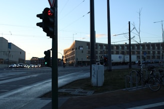 A city intersection with a traffic light showing red. Multiple vehicles, including a truck, are queued along the road. A modern building with many windows is visible in the background. There are bicycles parked near a utility pole, and the area appears quiet with few pedestrians visible. The sky is clear with shades of blue and orange as the sun sets.