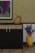 A modern interior scene featuring a cabinet with a sleek black and wood finish. On top of the cabinet, there is an abstract orange sculpture with a circular design and a flat decorative element. A framed abstract painting hangs on the wall above, displaying a variety of colors including purple, green, and blue. To the right of the cabinet, a geometric yellow vase holds dried branches, adding a touch of natural texture. The background features a textured yellow wallpaper, complementing the wooden floor.