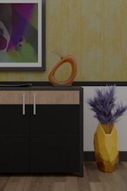 A modern interior scene featuring a cabinet with a sleek black and wood finish. On top of the cabinet, there is an abstract orange sculpture with a circular design and a flat decorative element. A framed abstract painting hangs on the wall above, displaying a variety of colors including purple, green, and blue. To the right of the cabinet, a geometric yellow vase holds dried branches, adding a touch of natural texture. The background features a textured yellow wallpaper, complementing the wooden floor.