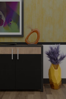 A modern interior scene featuring a cabinet with a sleek black and wood finish. On top of the cabinet, there is an abstract orange sculpture with a circular design and a flat decorative element. A framed abstract painting hangs on the wall above, displaying a variety of colors including purple, green, and blue. To the right of the cabinet, a geometric yellow vase holds dried branches, adding a touch of natural texture. The background features a textured yellow wallpaper, complementing the wooden floor.