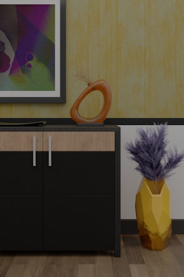 A modern interior scene featuring a cabinet with a sleek black and wood finish. On top of the cabinet, there is an abstract orange sculpture with a circular design and a flat decorative element. A framed abstract painting hangs on the wall above, displaying a variety of colors including purple, green, and blue. To the right of the cabinet, a geometric yellow vase holds dried branches, adding a touch of natural texture. The background features a textured yellow wallpaper, complementing the wooden floor.