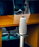 A modern office setting with sleek black office chairs around a wooden table. At the forefront, a tall, cylindrical speaker or device stands on a metallic stand.