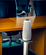 A modern office setting with sleek black office chairs around a wooden table. At the forefront, a tall, cylindrical speaker or device stands on a metallic stand.