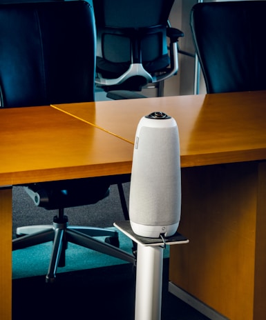 A modern office setting with sleek black office chairs around a wooden table. At the forefront, a tall, cylindrical speaker or device stands on a metallic stand.