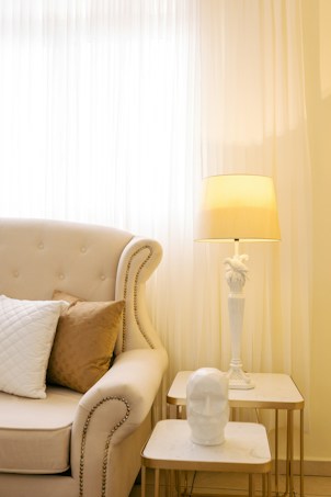 A cozy and elegant room corner featuring an upholstered armchair with rivet detailing, adorned with white and brown cushions. A small side table holds an artistic white bust, and a taller table supports a decorative white lamp with a bird design. The background is softly lit by sheer floor-length curtains, creating a warm and inviting atmosphere.