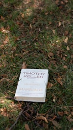 A book titled 'The Meaning of Marriage' by Timothy Keller is placed on the grass. The background consists of green and brown grass, with fallen leaves scattered around.