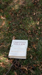 A book titled 'The Meaning of Marriage' by Timothy Keller is placed on the grass. The background consists of green and brown grass, with fallen leaves scattered around.