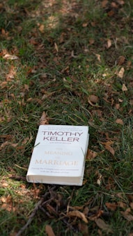 A book titled 'The Meaning of Marriage' by Timothy Keller is placed on the grass. The background consists of green and brown grass, with fallen leaves scattered around.