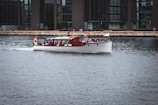 A classic wooden boat with a white hull cruises gently across a calm urban waterway, adorned with a red and white flag at the rear. The background features modern office buildings with large glass windows lining the waterfront. A few people can be seen along the waterfront enjoying the view.