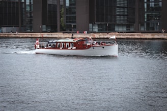 A classic wooden boat with a white hull cruises gently across a calm urban waterway, adorned with a red and white flag at the rear. The background features modern office buildings with large glass windows lining the waterfront. A few people can be seen along the waterfront enjoying the view.