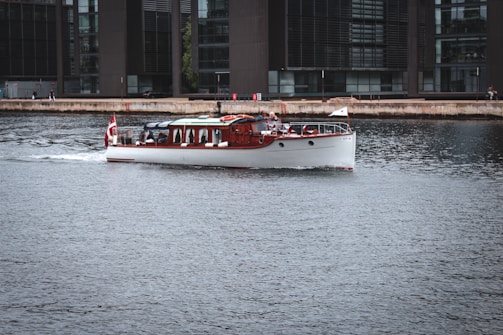 A classic wooden boat with a white hull cruises gently across a calm urban waterway, adorned with a red and white flag at the rear. The background features modern office buildings with large glass windows lining the waterfront. A few people can be seen along the waterfront enjoying the view.