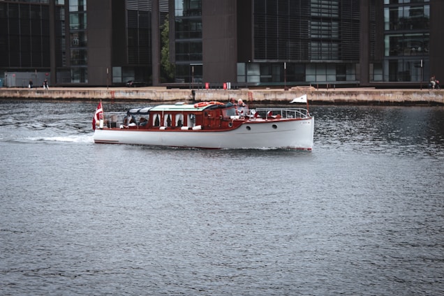 A classic wooden boat with a white hull cruises gently across a calm urban waterway, adorned with a red and white flag at the rear. The background features modern office buildings with large glass windows lining the waterfront. A few people can be seen along the waterfront enjoying the view.