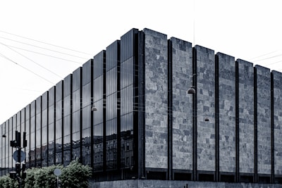 A modern building features a facade with vertical panels of glass and stone, creating a strong, geometric pattern. Overhead, several suspended streetlights and numerous electrical wires add a layer of complexity to the scene. Lush greenery is visible at the base of the building.