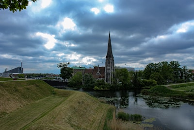 A serene landscape featuring a Gothic-style church with a tall spire surrounded by lush greenery. The church is reflected in a calm body of water in the foreground, with a grassy embankment leading up to it. The sky is overcast with dramatic clouds, adding a moody atmosphere to the scene. In the background, modern buildings are partially visible.