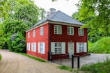 A charming two-story red house with white window frames is nestled in a lush, green garden. The structure features a grey-tiled roof and is surrounded by dense foliage with a variety of trees and plants. A gravel path curves gently around the house, leading to a small cobblestone area adjacent to the building.
