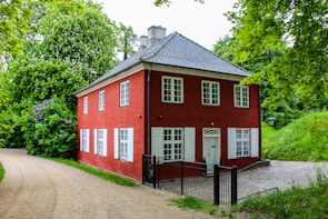 A charming two-story red house with white window frames is nestled in a lush, green garden. The structure features a grey-tiled roof and is surrounded by dense foliage with a variety of trees and plants. A gravel path curves gently around the house, leading to a small cobblestone area adjacent to the building.