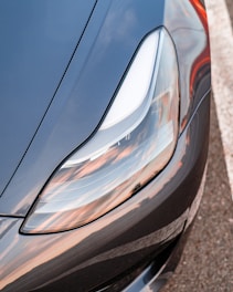 Close-up view of a sleek, modern car headlight with reflections of a sunset sky. The headlight design is streamlined and smooth, with a focus on precision and style. The car's exterior features a metallic finish, showcasing a mix of blue and silver tones.