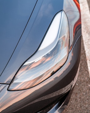 Close-up view of a sleek, modern car headlight with reflections of a sunset sky. The headlight design is streamlined and smooth, with a focus on precision and style. The car's exterior features a metallic finish, showcasing a mix of blue and silver tones.