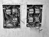The image depicts two electrical boxes mounted on a wall. Both boxes are open and show numerous wires and circuits in a chaotic arrangement. There is dirt and signs of wear and tear on the boxes and the surrounding wall, indicating they are old and possibly neglected.