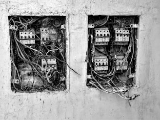 The image depicts two electrical boxes mounted on a wall. Both boxes are open and show numerous wires and circuits in a chaotic arrangement. There is dirt and signs of wear and tear on the boxes and the surrounding wall, indicating they are old and possibly neglected.